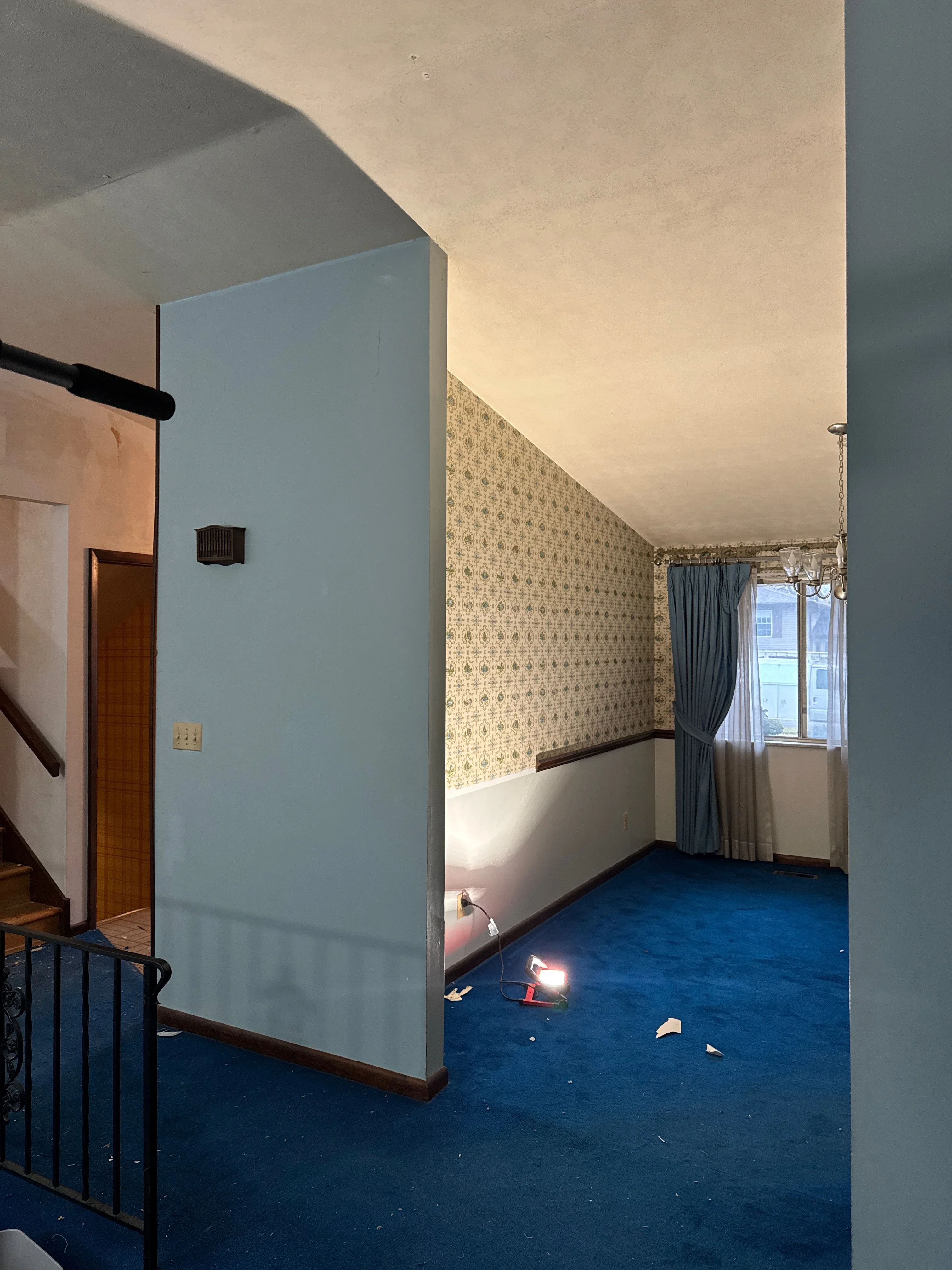 Interior room with light blue drywall partition, patterned wallpaper, blue carpet, and window with curtains